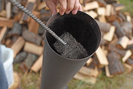 Technicien de ramonage portant vêtements de travail professionnels, utilisant une brosse hérisson et tiges de ramonage pour nettoyer l'intérieur d'un conduit de cheminée en pierre ou en brique depuis l'ouverture de foyer d'une cheminée traditionnelle, dans un intérieur résidentiel, mettant en évidence l'intervention technique d'entretien du système de chauffage au bois.